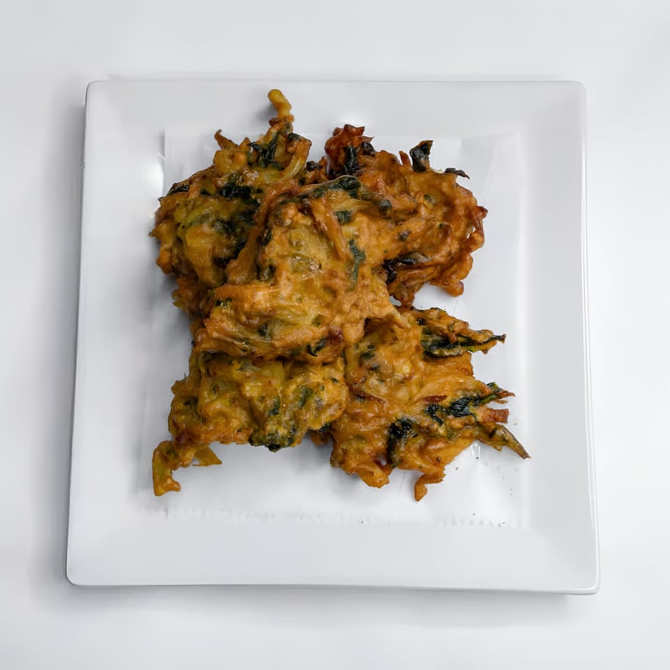 Golden-brown Indian pakora, a deep-fried fritter with visible green herbs, served on a white square plate.