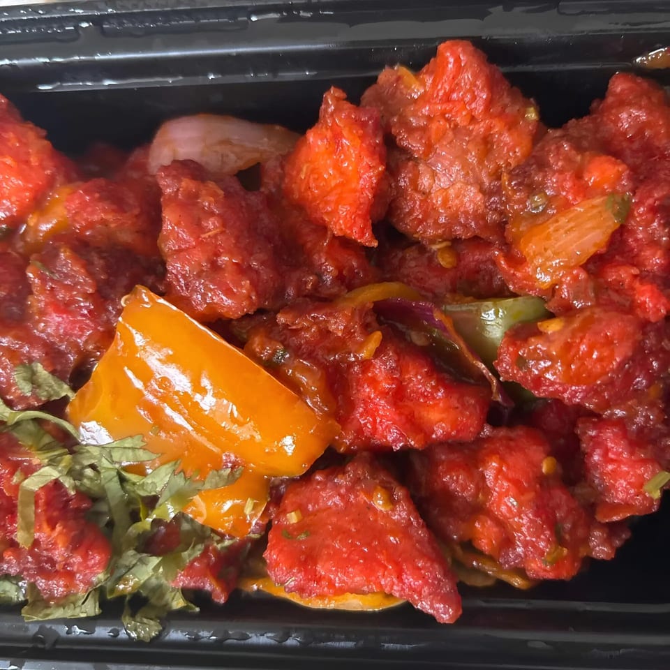 Indian Aroma's vibrant red chili chicken with bell peppers and herbs in a black container.